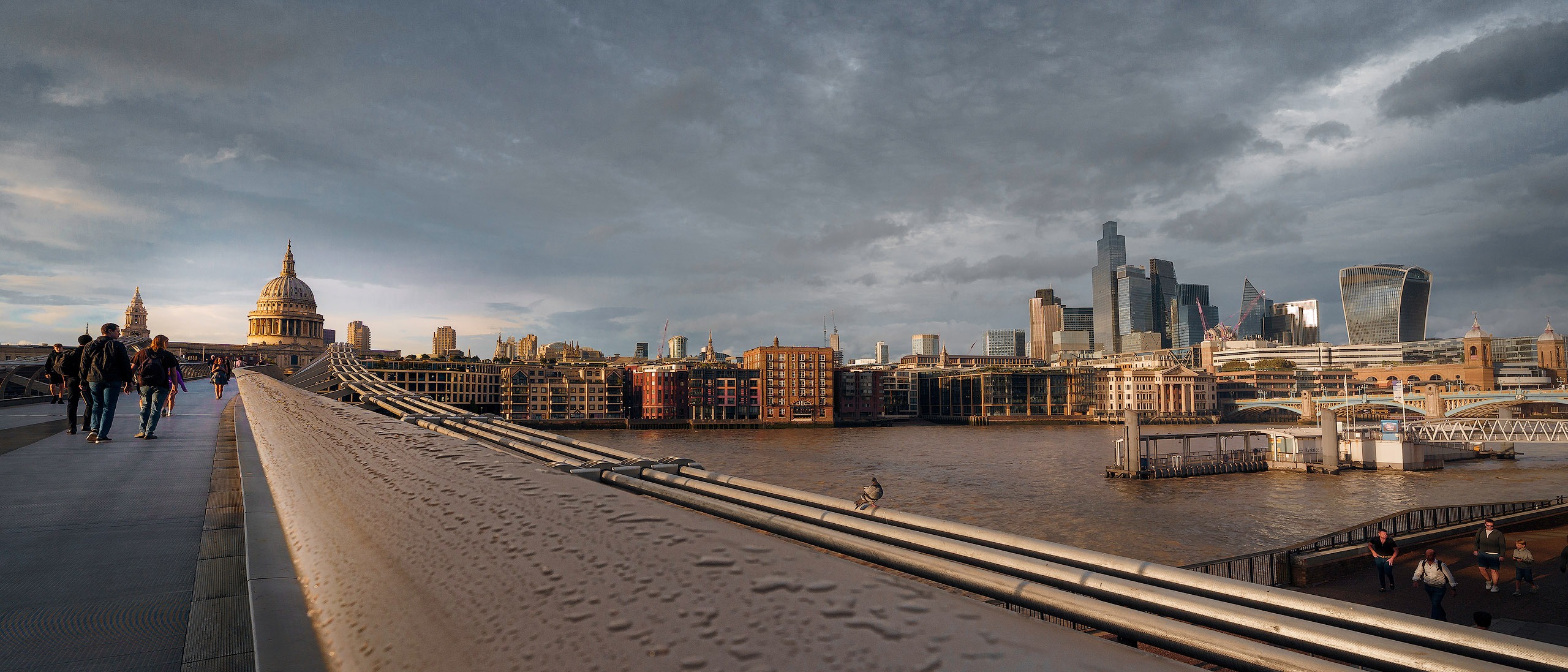 Fine-Art-Fotografie: Weitwinkelblick über die Millennium Bridge am Abend, belebt durch vereinzelte Fußgänger. Die stählerne Konstruktion führt den Blick diagonal zur links positionierten St. Paul’s Cathedral. Rechts ist das beleuchtete Themseufer mit Gebäuden zu sehen.