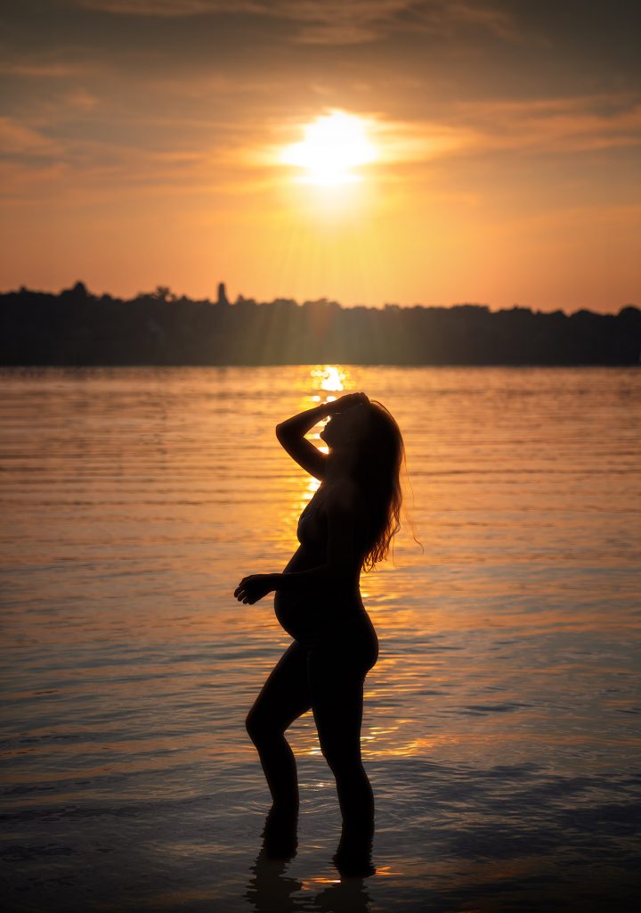 Stimmungsvolles Porträt im Gegenlicht: Eine schlanke, hochschwangere Frau steht als dunkle Silhouette im seichten Wasser eines Sees bei Sonnenuntergang. Ihr deutlich sichtbarer Babybauch zeichnet sich klar ab, während sie den Kopf nach hinten geworfen hat und einen Arm auf ihre Stirn legt. Im Hintergrund ist das andere Ufer mit den dunklen Silhouetten von Bäumen zu sehen; die untergehende Sonne taucht den Himmel und das Wasser in eine warme, stimmungsvolle Abendbeleuchtung.