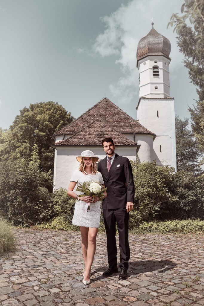 Ein junges Brautpaar vor einer kleinen weißen Kirche auf einem gepflasterten Weg. Der Bräutigam trägt einen Anzug, die Braut ein kurzes Spitzenkleid sowie einen Hut und hält einen Blumenstrauß in der Hand. Das Wetter ist schön bei einem klaren, blauen Himmel.