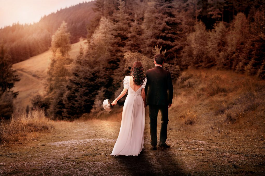 Rückenansicht eines Brautpaars, das in der letzten Abendsonne Hand in Hand einen Weg entlanggeht. Die Braut trägt ein langes, weißes Kleid mit Puffärmeln und hat lange, dunkelbraune Locken. Der Bräutigam trägt einen Smoking und hat dunkles Haar. Im Hintergrund ist ein Wald zu sehen. Das Foto ist komplett in warmes, stimmungsvolles Abendlicht getaucht.