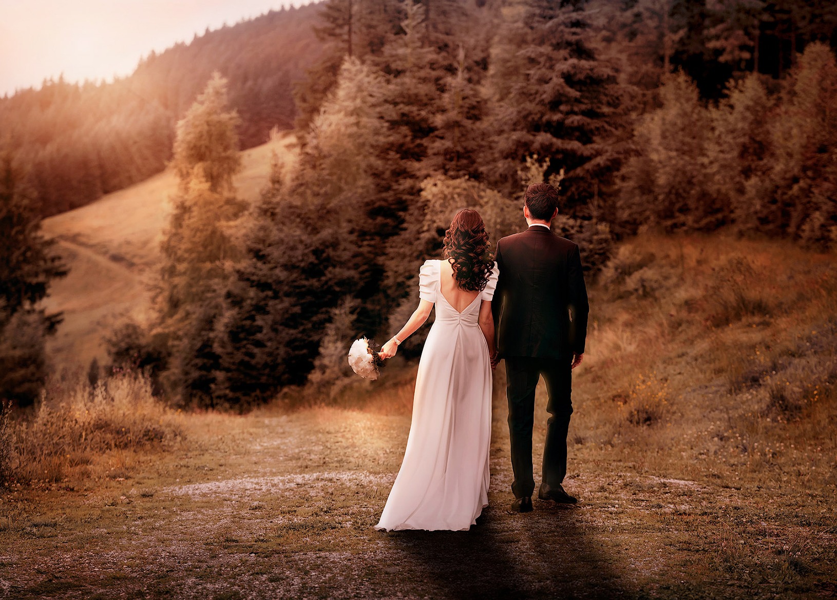Rückenansicht eines Brautpaars, das in der letzten Abendsonne Hand in Hand einen Weg entlanggeht. Die Braut trägt ein langes, weißes Kleid mit Puffärmeln und hat lange, dunkelbraune Locken. Der Bräutigam trägt einen Smoking und hat dunkles Haar. Im Hintergrund ist ein Wald zu sehen. Das Foto ist komplett in warmes, stimmungsvolles Abendlicht getaucht.
