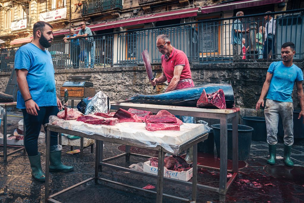 Eine Straßenszene am Marktstand in Catania. Drei Männer stehen um einen Markttisch, auf dem große Thunfischstücke ausgebreitet sind. Der eine Fischverkäufer hält ein großes Beil in der Hand. Im Hintergrund begrenzt eine Mauer eine höher gelegte Straße, auf der Passanten und Wohnhäuser zu sehen sind.