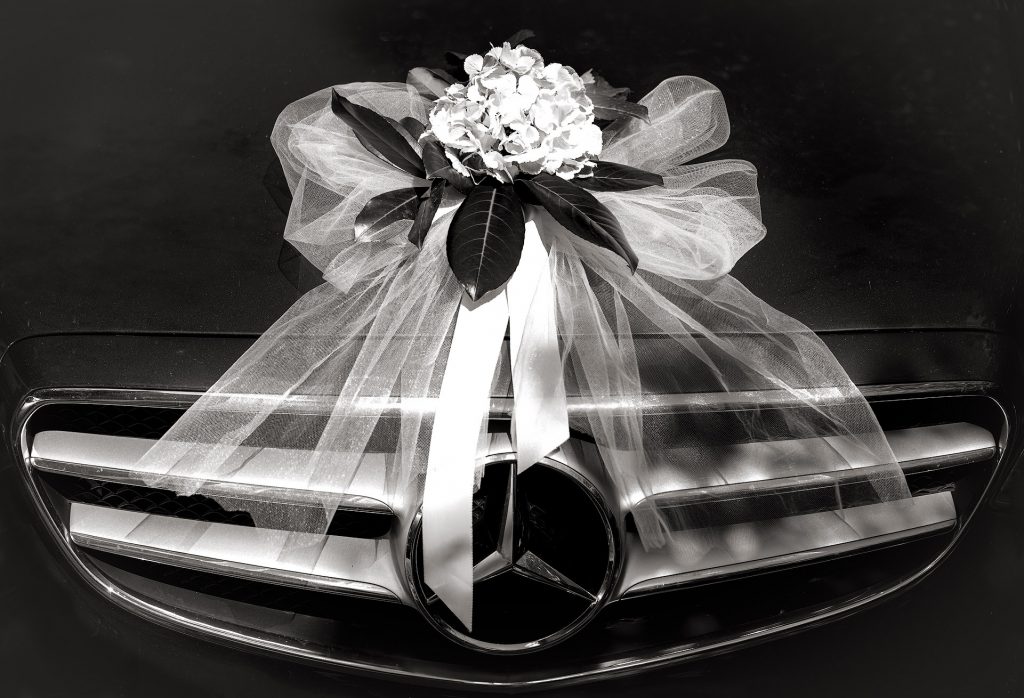 Schwarz-Weiß-Fotografie: Frontansicht eines dunklen Mercedes-Autos mit festlicher Hochzeitsdekoration auf der Motorhaube und dem Kühler. Darauf befindet sich eine sehr große, weiße Tüllschleife, auf der eine weitere kleiner Schleife und eine weiße Hortensie platziert sind.