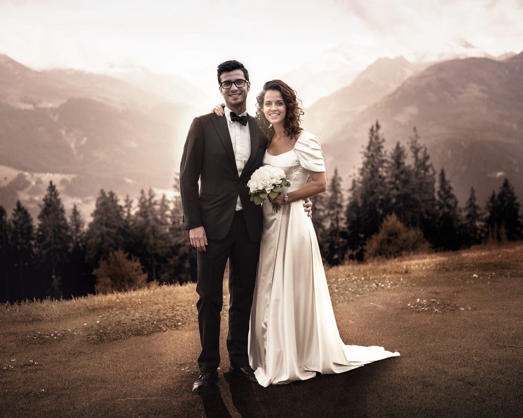Hochzeitsfotografie eines dunkelhaarigen Brautpaares vor einer beeindruckenden alpinen Bergkulisse: Die Braut im eleganten langen weißen Hochzeitskleid und der Bräutigam mit Brille im klassischen Smoking posieren vor dem Panorama der Alpen bei Sonnenuntergang. Die ganze Bild ist in warmes Abendlicht getaucht.