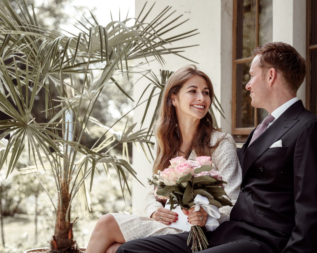 Ein junges Hochzeitspaar auf einer Bank. Sie strahlt ihn verliebt und er schaut ihr in die Augen. In den Händen hält sie einen Rosenstrauß. Im Hintergrund eine Palme.