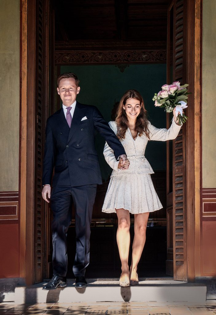 Emotionaler Hochzeitsmoment: Das Brautpaar verlässt Hand in Hand das Standesamt, während die Braut glücklich den Blumenstrauß in die Luft reckt; der dunkle Hintergrund im Türrahmen lässt das Paar strahlen.