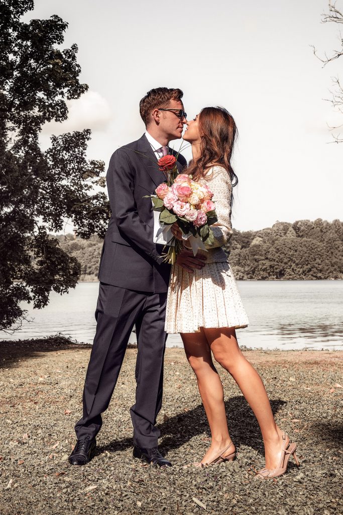 Hochzeitsfotografie am Seeufer: Ein frisch vermähltes Hochzeitspaar im modernen Standesamt-Outfit küsst sich innig vor der malerischen Kulisse des ruhigen Wassers.