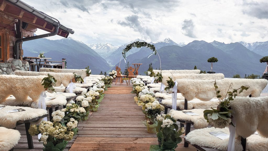 Eventfotografie einer alpinen Outdoor-Hochzeitslocation vor majestätischer Bergkulisse: Eine Allee aus hölzernen Bänken, dekoriert mit weißen Hortensien und gemütlichen Schaffellen, führt perspektivisch auf einen prachtvollen Hochzeitsbogen zu.