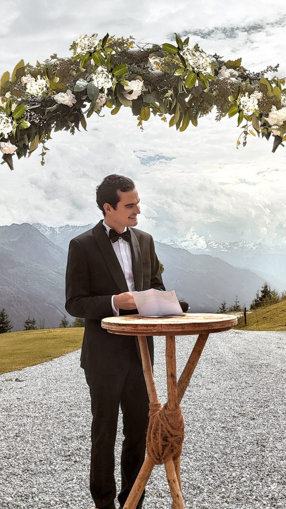 Ein junger Mann im Smoking vor einer beeindruckenden Bergkulisse. Er steht an einem Stehpult und hält ein Schriftstück, für seine Hochzeitsrede in der Hand. Über ihm eine Blumengirlande mit weißen Blüten.