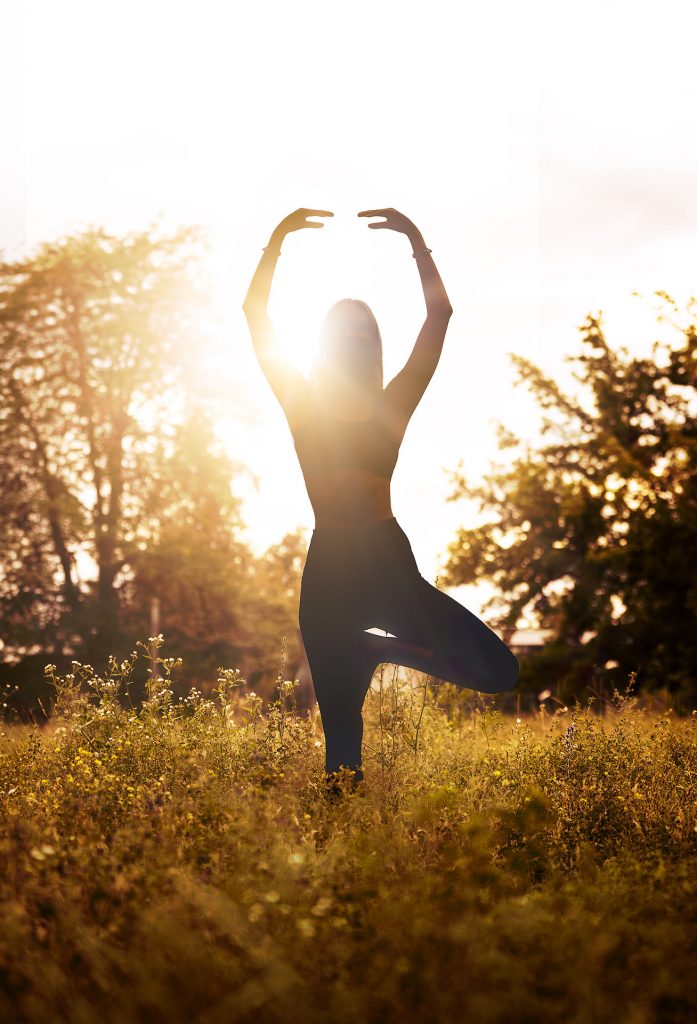 Sehr atmosphärische Fine-Art-Aufnahme der Yogalehrerin Lena als dunkle Silhouette. Sie steht in der Pose des Yogabaums auf einer Wiese im direkten Gegenlicht der untergehenden Sonne, die den Himmel erleuchtet. Ihr Umriss wird vom Licht gezeichnet, die Hände sind über dem Kopf gestreckt.