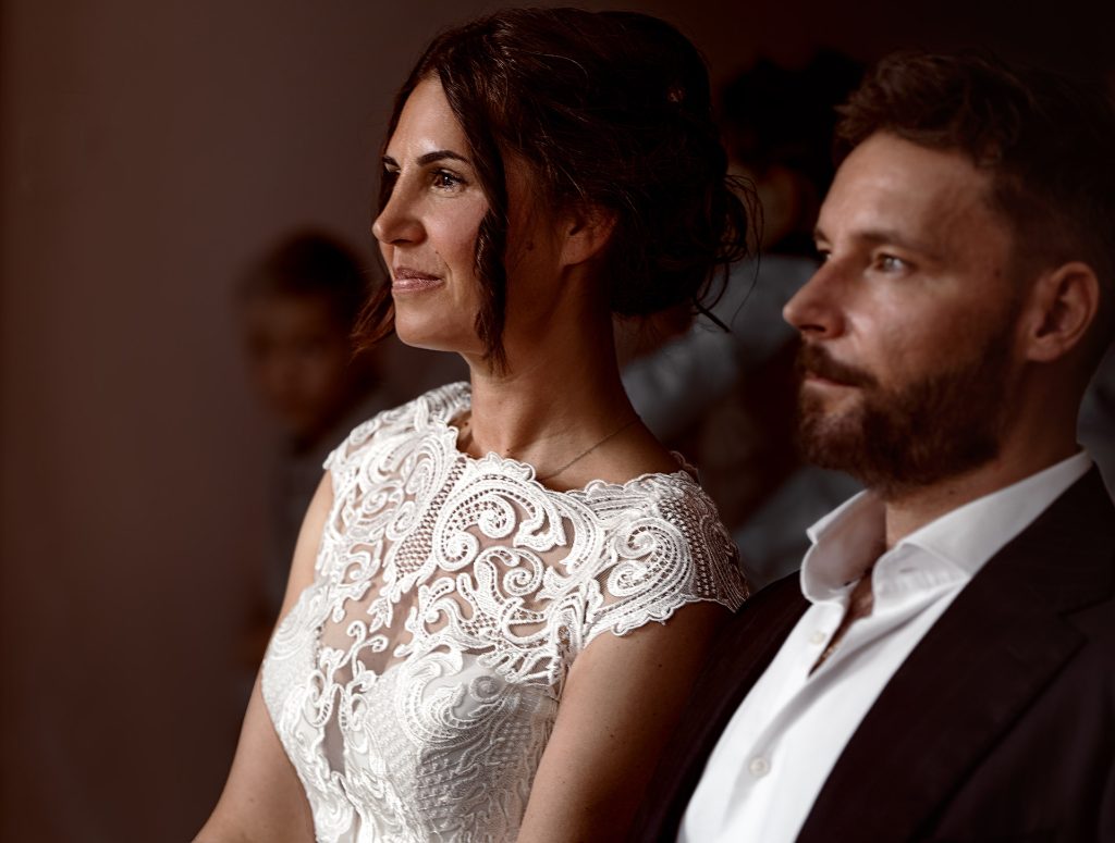 Hochzeitspaar, seitliches Portrait bis Brust, Fokus auf Braut in weißem Spitzenoberteil mit hochgesteckten Haaren, er sitzt vor ihr und ist außer Fokus, Hintergrund dunkel und ebenfalls verschwommen.