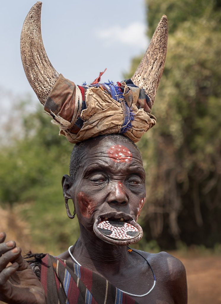 Porträt einer sehr alten Mursi-Frau. Ihre Augen sind blind, sie trägt einen bemalten Lippenteller aus Ton im Mund und hat rote Ockerkreise auf das Gesicht gemalt. Die geschlitzten Ohrläppchen hängen als weite Schlaufen herab. Auf dem Kopf trägt sie einen großen Kopfputz aus zwei abstehenden Bullenhörnern, die in der Mitte mit bunten Stofflumpen umwickelt sind. Der Hintergrund ist verschwommen.