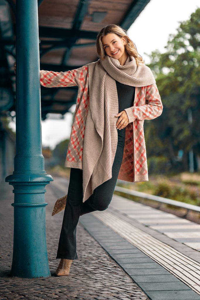 Mode-Shooting am Bahnhof: Blondes Modell mit langen Locken posiert an einer Säule auf dem Bahnsteig. Sie trägt einen karierten Strickmantel und einen groben Strickschal vor einem natürlichen Hintergrund mit Bäumen.