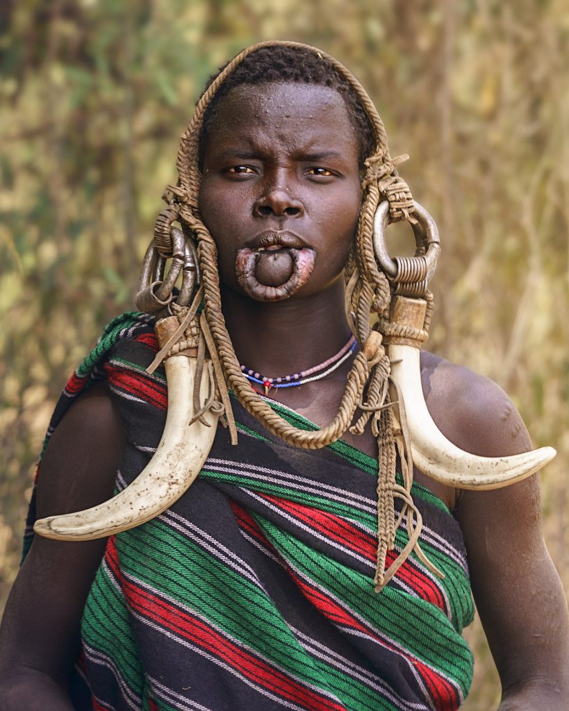 Porträt einer jungen Mursi-Frau, die frontal in die Kamera blickt. Sie trägt einen aufwendigen Kopfschmuck aus Warzenschwein-Hauern, die an schweren Metallringen und Lederbändern befestigt sind. Ihr Körper ist in ein traditionell gewebtes Tuch gehüllt, das schwarz, rot und grün gestreift ist. Ihre Lippe ist für einen Lippenteller geschlitzt, der jedoch nicht eingesetzt ist.
