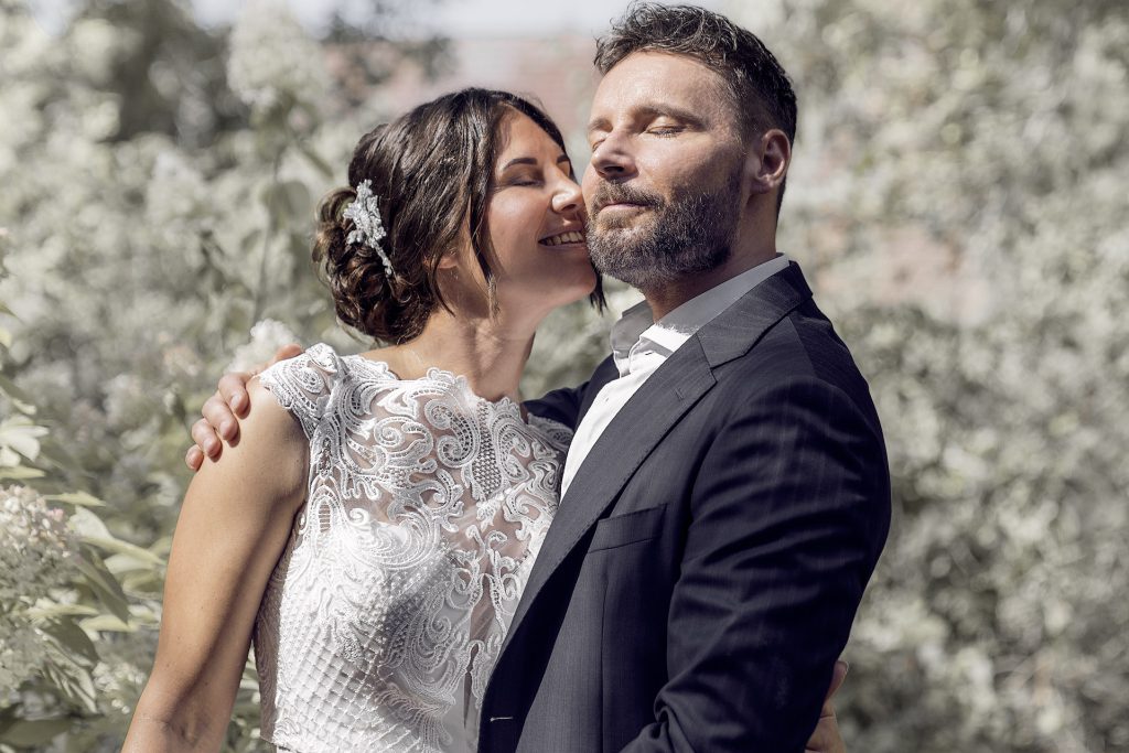 Ein Brautpaar, sie im weißen, ärmellosen Spitzenkleid bis zur Taille und hochgesteckten Haaren, er im dunklen Anzug mit weißem Hemd und Bart. Die Braut küsst den Bräutigam auf die Wange, während er die Augen geschlossen hält. Seine hintere Hand liegt auf ihrer Schulter. Der Hintergrund mit Blättern und Pflanzen ist hell.