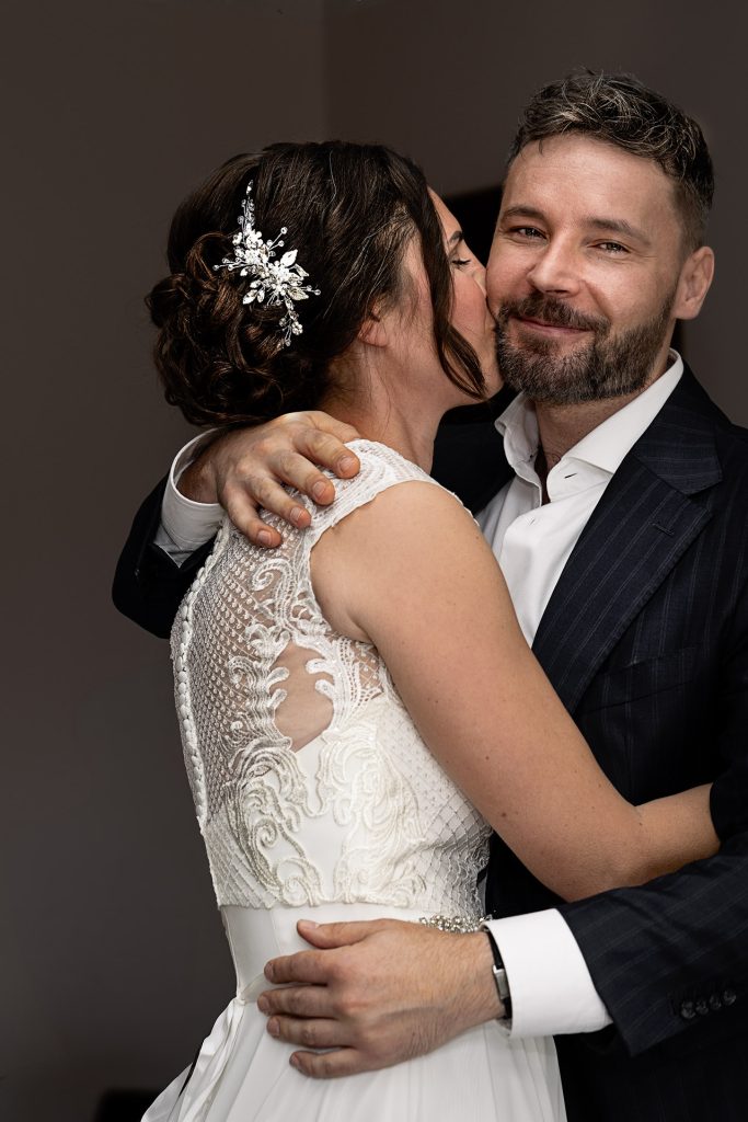 Ein Brautpaar, sie im weißen, ärmellosen Spitzenkleid bis zur Taille und hochgesteckten Haaren, er im dunklen Anzug mit weißem Hemd und Bart. Die Braut küsst den Bräutigam auf die Wange, während er lächelt. Seine vordere Hand liegt um ihre Taille, die andere auf ihrer Schulter. Der Hintergrund ist dunkel.