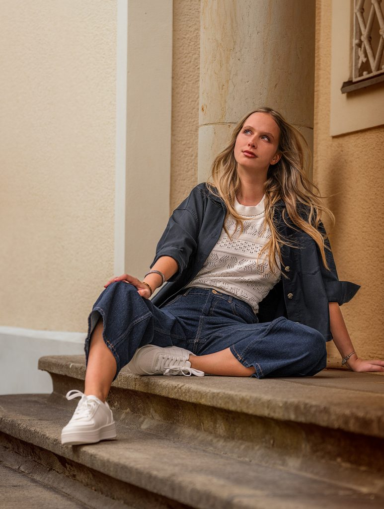 Modefoto in warmem, goldenem Licht: Ein langhaariges, blondes Model sitzt am Ende einer Treppe auf dem Fußboden vor einer gelben Wand und einer Säule. Ein Bein ist lang ausgestreckt, das andere angewinkelt. Sie trägt eine weite Jeans, eine dunkelblaue sportliche Jacke, einen weißen Häkelpulli sowie weiße Turnschuhe und blickt verträumt zum oberen linken Bildrand in das einfallende Sonnenlicht.