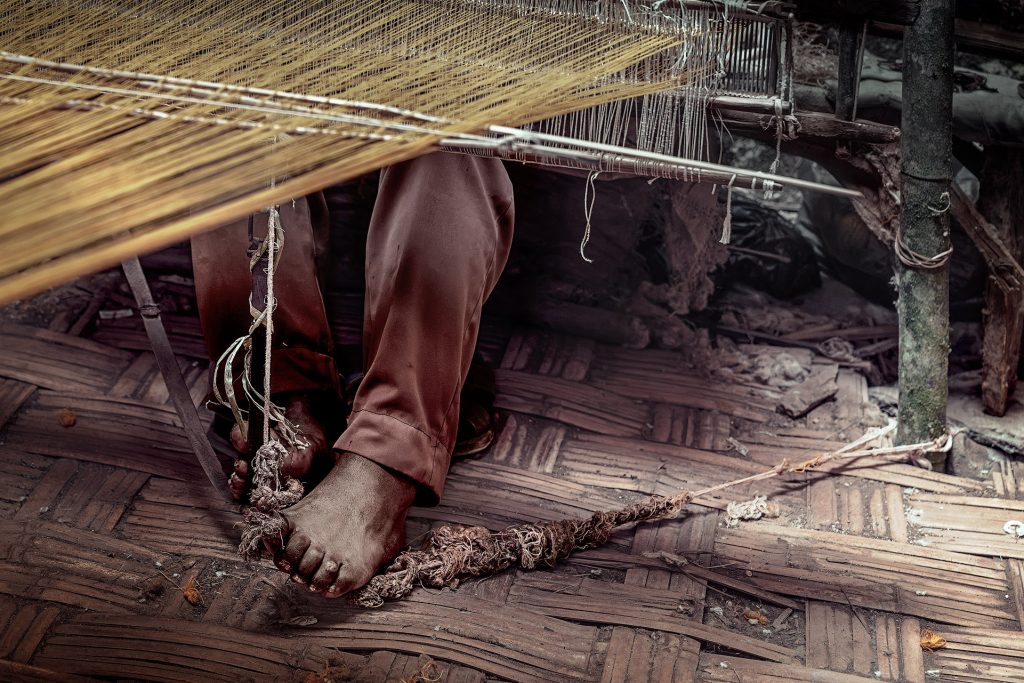 Detailaufnahme des Dorze-Webers. Die Kettfäden verlaufen diagonal von der oberen Mitte nach links unten durch das Bild. Unterhalb der Webmechanik arbeiten die Beine des Webers mit einem unbeschuhten Fuß, der auf einem einfach, grob geflochtenen Schilfteppich ruht.