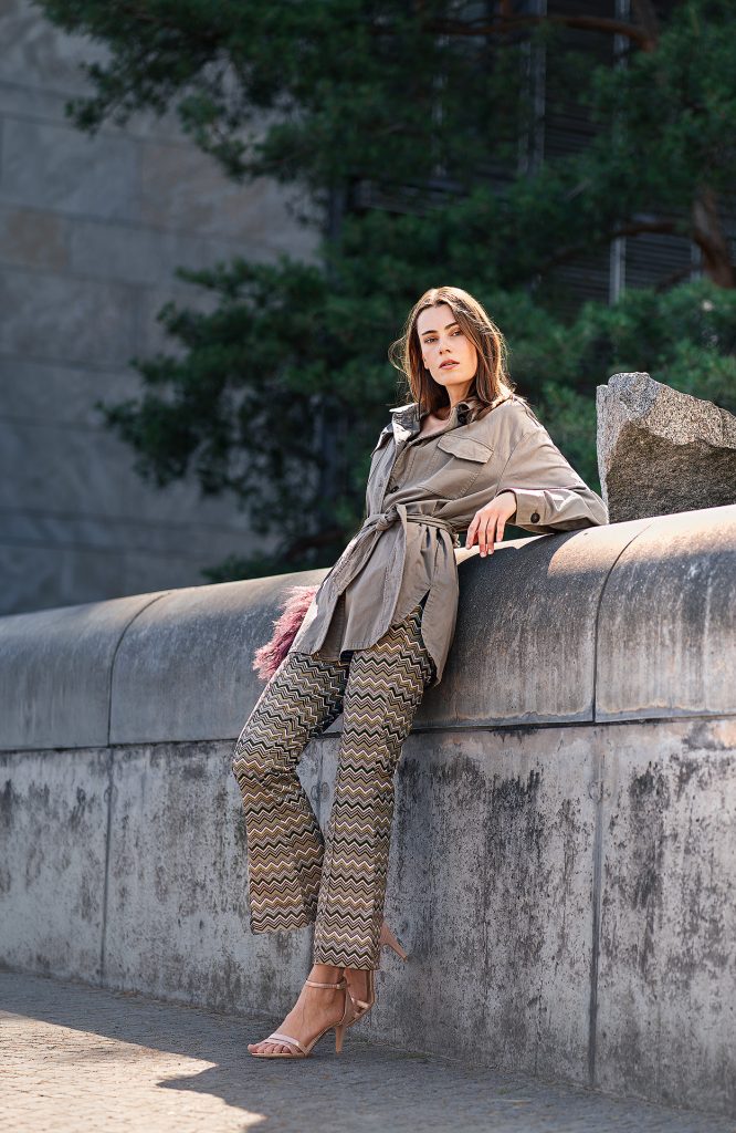 Urbanes Mode-Shooting: Modell in einer auffälligen Hose mit grafischem Muster und sportlicher Jacke lehnt lässig an einer Betonmauer vor städtischer Kulisse.