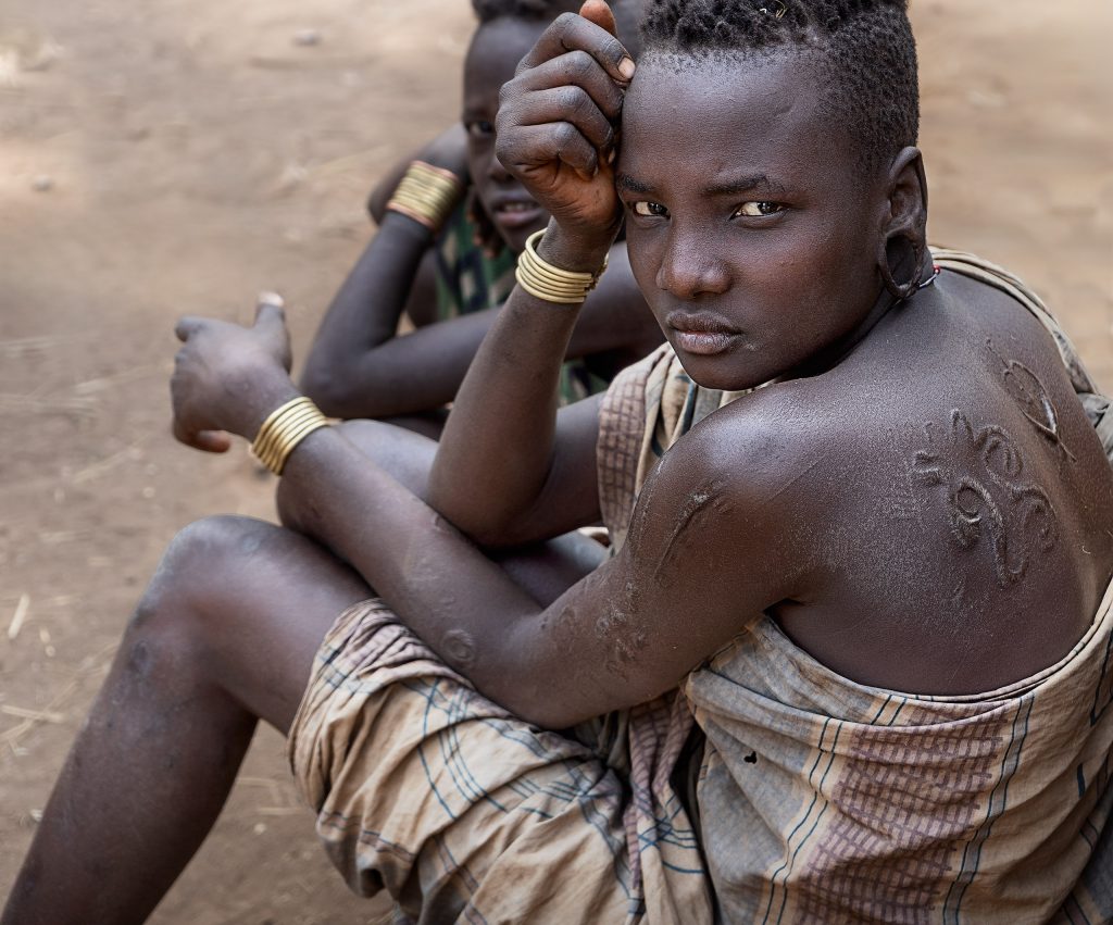 Eine von oben fotografierte Aufnahme einer jungen Mursi-Frau im gelben Sand. Sie sitzt auf dem Boden und ist halb seitlich, halb von rückwärts zu sehen, während sie den Kopf nach oben zur Kamera dreht. Ihre Schultern und ihr Rücken sind mit kunstvollen Schmucknarben verziert. Als Bekleidung trägt sie lediglich ein Tuch, das auf der abgewandten Schulter geknotet ist. Im Hintergrund ist verschwommen ein weiteres Mursi-Mädchen zu erkennen.