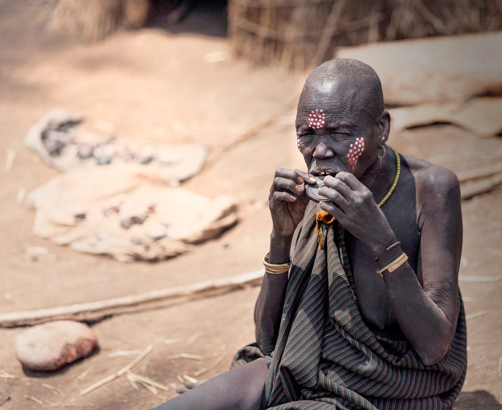 Mursi Inserting her Lip Plate Foto einer kahlköpfigen alten Mursi-Frau mit geschlossenen Augen. Sie trägt die typische Perlhuhn-Kreisbemalung auf Wange und Stirn, bestehend aus einem großen roten Fleck mit kleinen weißen Punkten. Mit beiden Händen hält sie einen Lippenteller und setzt diesen gerade in ihre geschlitzte Unterlippe ein. Sie sitzt auf dem Fußboden im Hintergrund ist undeutlich eine Strohhütte zu erkennen.