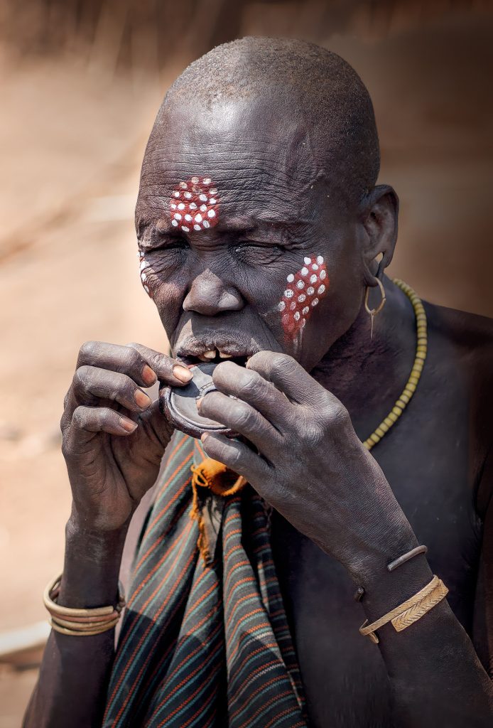 Porträt einer kahlköpfigen alten Mursi-Frau mit geschlossenen Augen. Sie trägt die typische Perlhuhn-Kreisbemalung auf Wange und Stirn, bestehend aus einem großen roten Fleck mit kleinen weißen Punkten. Mit beiden Händen hält sie einen Lippenteller und setzt diesen gerade in ihre geschlitzte Unterlippe ein.