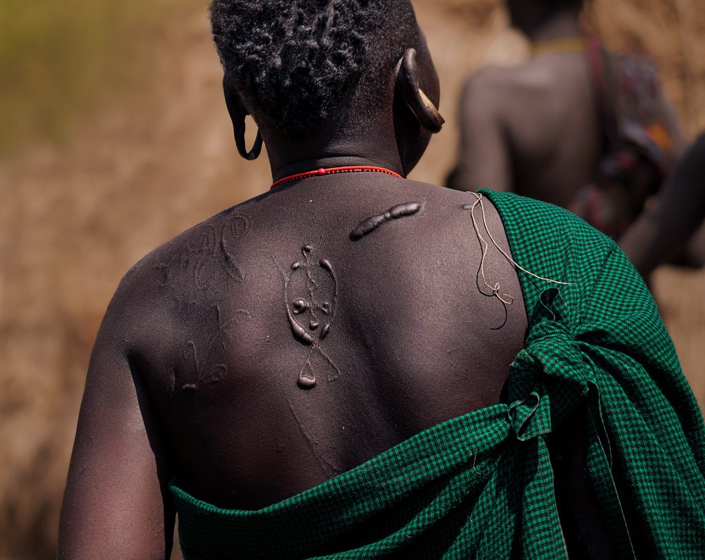 Rückansicht einer Mursi-Frau. Über einer Schulter trägt sie ein grün-schwarzes Hahnentritt-Tuch, das mit einem Knoten befestigt ist; der Rücken auf der anderen Seite ist frei und zeigt ornamentale Schmucknarben. Ein Ohr hat einen Holzpflock, während das andere Ohrläppchen geschlitzt ist und herabhängt. Der Hintergrund ist verschwommen in Gelb- und Orangetönen gehalten, und rechts vor der Frau laufen zwei weitere, verschwommene Mursi.