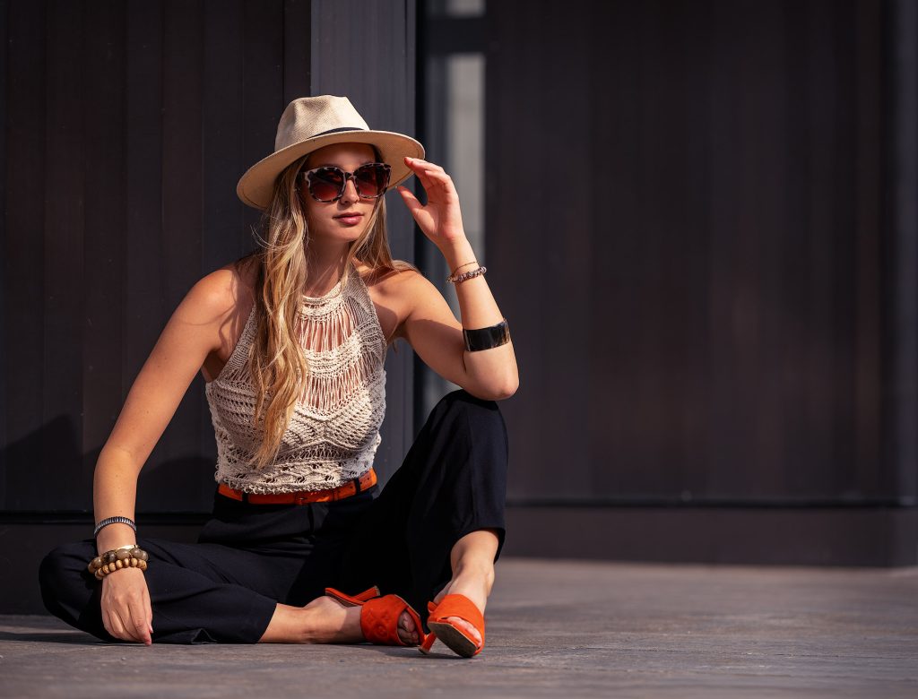 Ein langhaariges, blondes Model sitzt vor dunklen Wänden auf dem Boden; ein Bein ist angewinkelt, das andere seitlich lang ausgestreckt. Sie trägt eine schwarze Hose mit einem orangen Gürtel, ein beiges Häckeltop, orange Pantoletten, einen Panamahut sowie eine große Sonnenbrille. Eine Hand greift an den Hut.