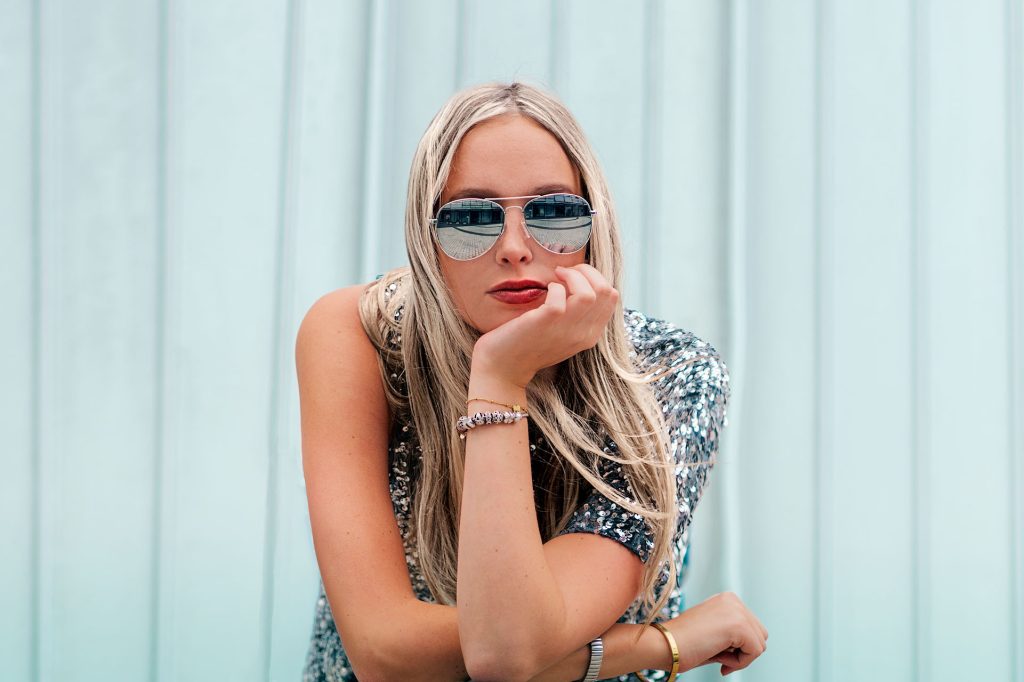 Ein hellblondes, langhaariges Model blickt herausfordernd direkt in die Kamera. Ihr Kinn ruht auf ihrer Hand, während die Ellbogen auf einer nicht sichtbaren Unterlage abgestützt sind und nur dieser Teil des Oberkörpers zu sehen ist. Sie trägt ein silbernes Paillettentop mit einer freien Schulter sowie eine verspiegelte Piloten-Sonnenbrille. Der Hintergrund ist sehr hell und mit vertikalen Längsstreifen versehen.