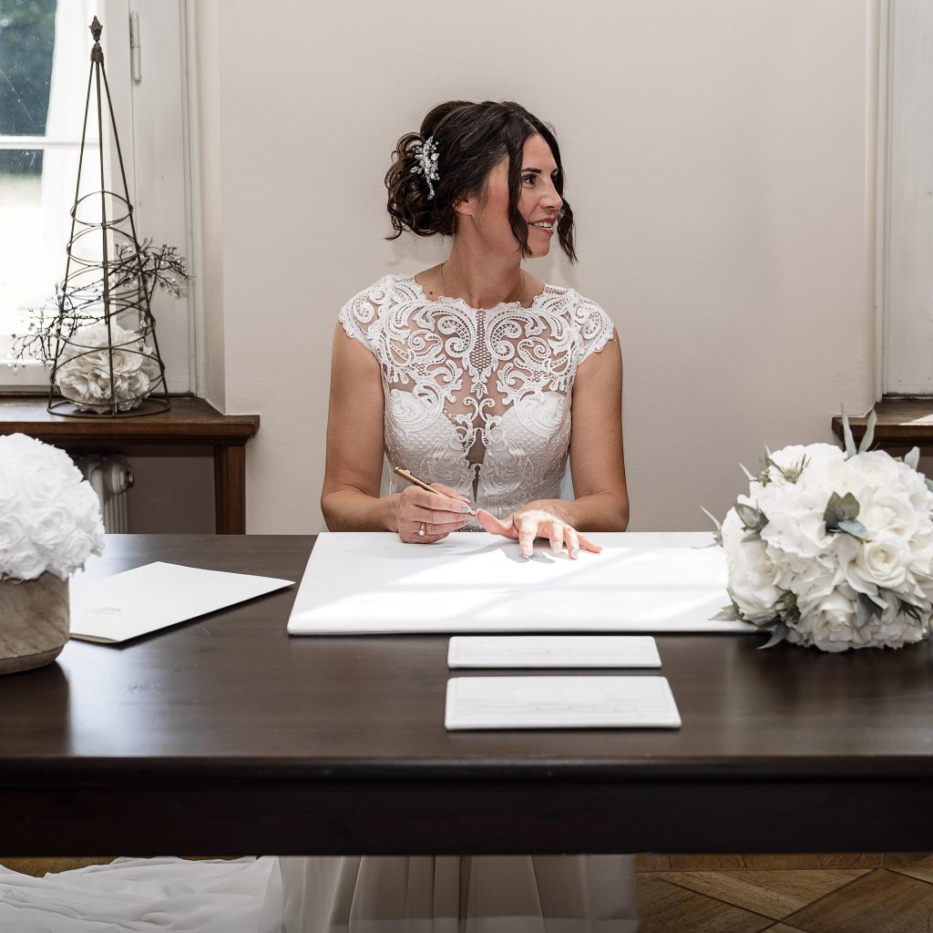 Braut beim Standesamt mit dunklem, hochgestecktem Haar in weißem Kleid mit armlosem Spitzenoberteil. Sie sitzt hinter einem dunklen Tisch und hält einen Stift in der Hand, anscheinend ist sie bei der Unterschrift der Hochzeitsurkunde. Auf dem Tisch stehen links weiße Blumen, rechts liegt ihr Strauß. Sie schaut seitlich.