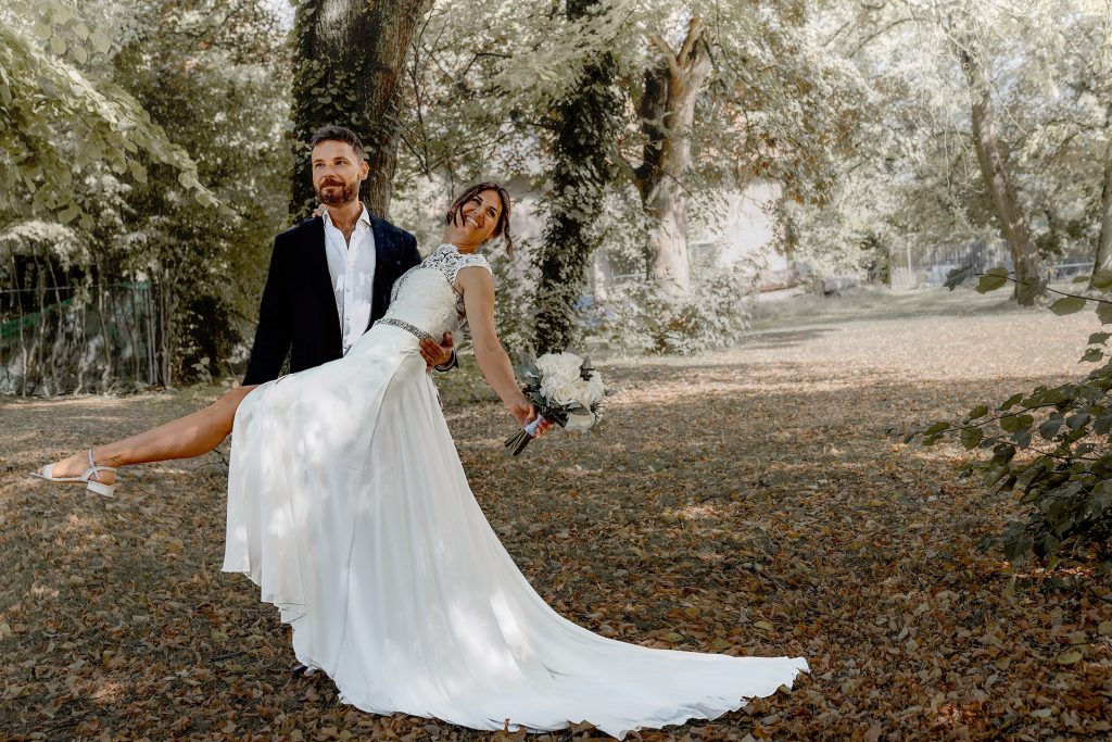 Hochzeitspaar in einem Park vor einem Baum. Er hat sie hochgehoben, wobei ihre lange Schleppe einen großen Teil unten im Bild bildet. Sie trägt ein weißes Kleid und dunkelhaariges, hochgestecktes Haar; er trägt einen dunklen Anzug. Ihre vordere Hand ist nach unten ausgestreckt und hält die Blumen, ihre andere Hand liegt auf seiner Schulter, und ein Bein ist waagerecht zum linken Bildrand ausgestreckt.
