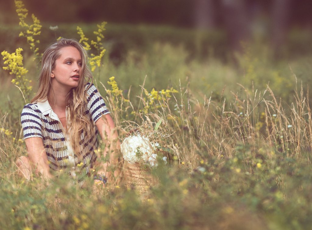 Ein Modefoto in einer Wiese mit Wildblumen und einem unscharfen Baumhintergrund: Ein langhaariges, blondes Model sitzt in der Wiese, wobei nur ihr Oberkörper im hohen Gras und den Blumen sichtbar ist. Sie trägt ein kurzes, blau-weiß gestreiftes Häkeltop und schaut zur Seite.